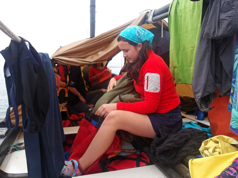A young woman wearing a red sweater and a blue bandana sits on a red bag inside a boat. She is looking down and appears to be organizing or packing her belongings. The boat is covered with a beige tarp, and various items of clothing are hanging around the interior. Other people are visible in the background, also seated inside the boat.